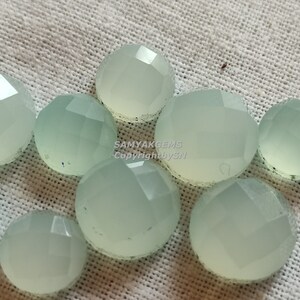 May include: A collection of ten pale green, faceted, round gemstone cabochons. The cabochons are arranged in a cluster on a white fabric background.