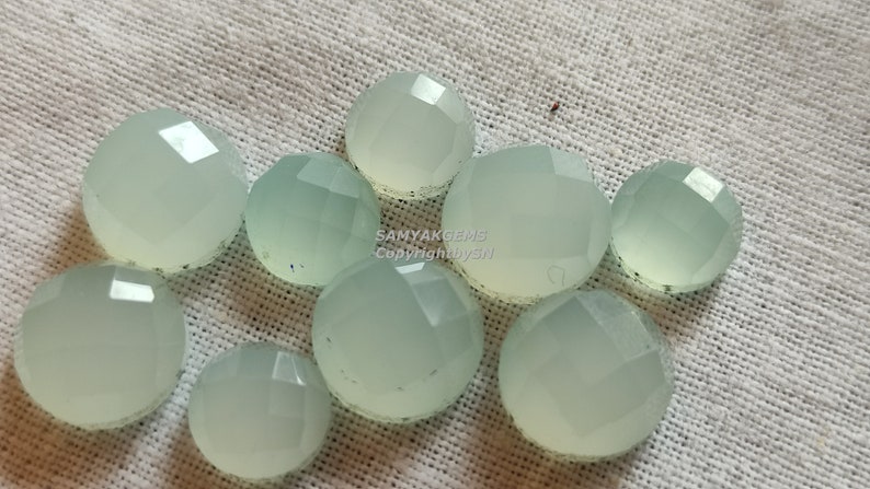 May include: A collection of ten pale green, faceted, round gemstone cabochons. The cabochons are arranged in a cluster on a white fabric background.