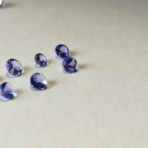 May include: A group of eight small, round, faceted gemstones in various shades of purple and blue. The gemstones are scattered on a white surface.