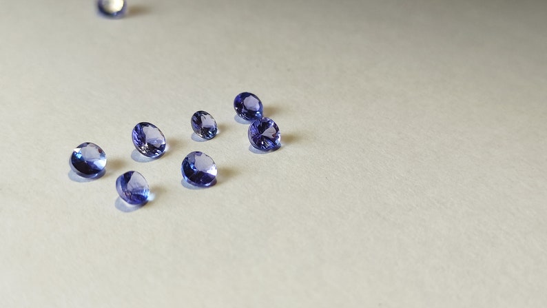 May include: A group of eight small, round, faceted gemstones in various shades of purple and blue. The gemstones are scattered on a white surface.