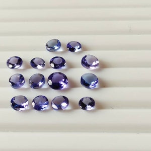 May include: A collection of 15 round, purple gemstones. The gemstones are various sizes and are arranged in a semi-circle on a white surface.