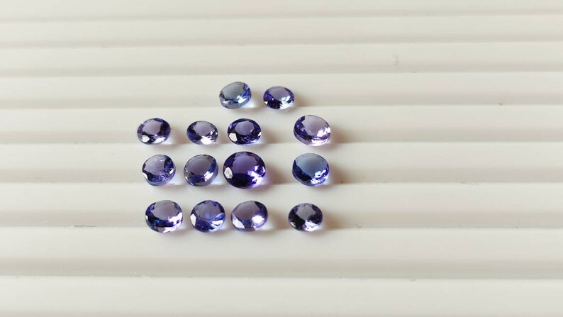 May include: A collection of 15 round, purple gemstones. The gemstones are various sizes and are arranged in a semi-circle on a white surface.