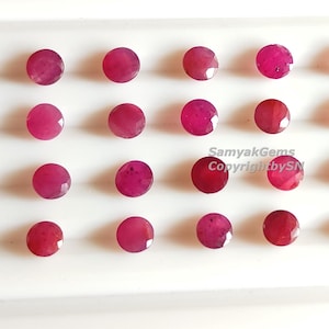 Könnte beinhalten: Eine Sammlung runder, facettierter Rubin-Edelsteine in verschiedenen Rot- und Rosatönen. Die Edelsteine sind in Reihen auf einer weißen Oberfläche angeordnet. Der Text "SamyakGems CopyrightbySN" ist enthalten.