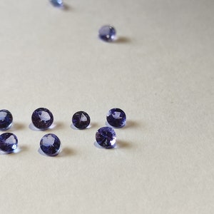 May include: A group of eight small, round, purple gemstones on a white surface.