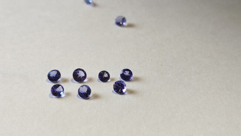 May include: A group of eight small, round, purple gemstones on a white surface.