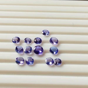 May include: A collection of round, purple gemstones on a white surface. The gemstones are of various sizes and have a slight sparkle.