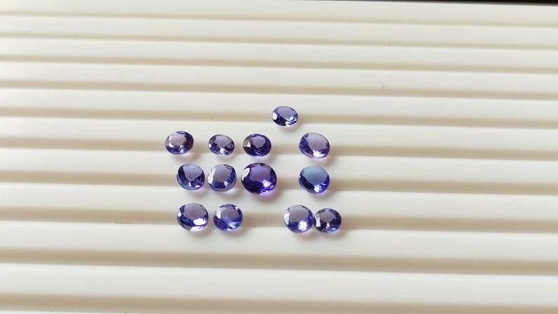 May include: A collection of round, purple gemstones on a white surface. The gemstones are of various sizes and have a slight sparkle.