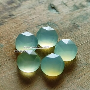 May include: Five faceted, light green gemstone beads on a wooden surface. The beads are round and have a slightly translucent appearance.