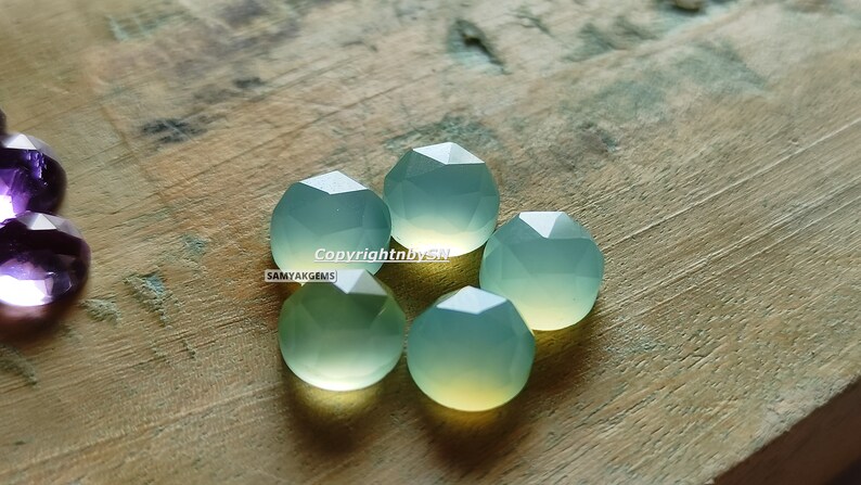May include: Five faceted, light green gemstone beads on a wooden surface. The beads are round and have a slightly translucent appearance.