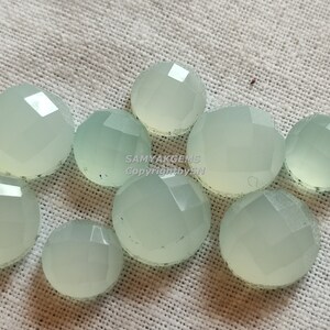 May include: A group of ten light green, faceted, round cabochon gemstones. The gemstones are arranged in a cluster on a white fabric background.