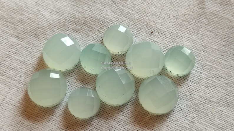 May include: A group of ten light green, faceted, round cabochon gemstones. The gemstones are arranged in a cluster on a white fabric background.