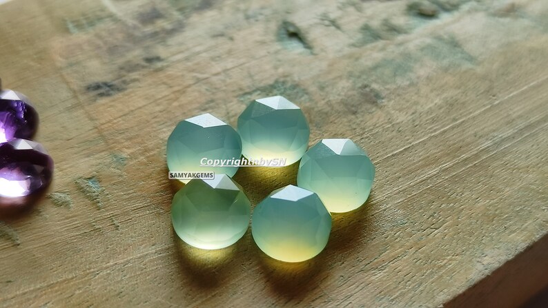 May include: Five faceted green chalcedony gemstones on a wooden surface. The gemstones are a light green colour with a slight yellow hue. The text "Copyright by SN SAMYAKGEMS" is visible in the image.