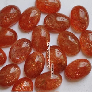 May include: A collection of 18 oval-shaped sunstone cabochons in shades of orange and red. The stones have a shimmery, iridescent quality.