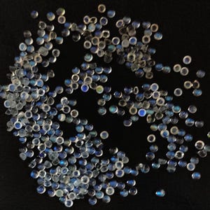 May include: A large group of iridescent blue and white gemstone cabochons, scattered on a black surface. The cabochons are round and have a smooth, polished finish.