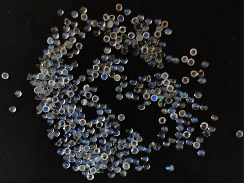 May include: A large group of iridescent blue and white gemstone cabochons, scattered on a black surface. The cabochons are round and have a smooth, polished finish.