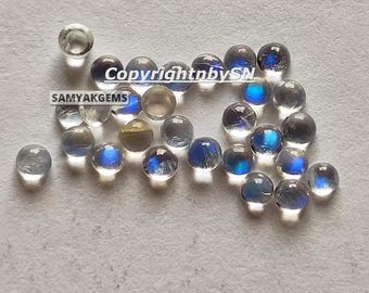May include: A collection of small, round, translucent cabochon gemstones with iridescent blue flashes. The stones are scattered, displaying varying levels of clarity and light reflection. Text overlay reads CopyrightnbySN and SAMYAKGEMS.