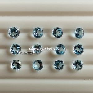 May include: Twelve round, faceted, light blue gemstones with a star-like pattern inside. The gemstones are arranged in three rows of four on a white surface.