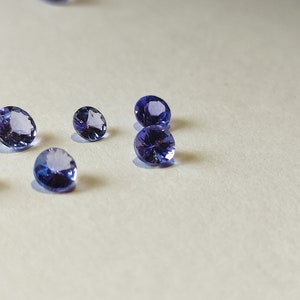 May include: A group of seven round, faceted, purple gemstones on a white surface.