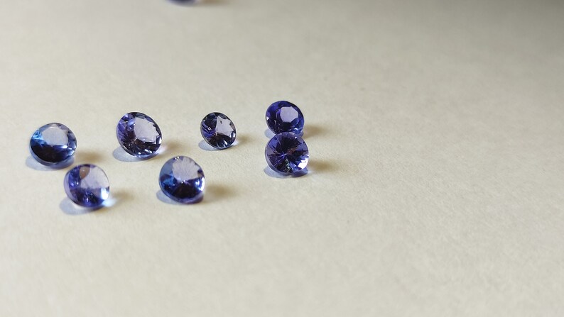 May include: A group of seven round, faceted, purple gemstones on a white surface.