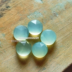 May include: Five light blue, faceted, round gemstone cabochons on a wooden surface. Copyright by SN SAMYAKGEMS.