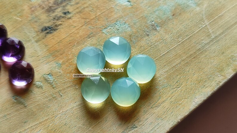 May include: Five light blue, faceted, round gemstone cabochons on a wooden surface. Copyright by SN SAMYAKGEMS.