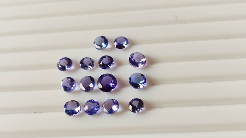 May include: A collection of round, purple gemstones, likely amethyst or topaz, arranged on a white surface.