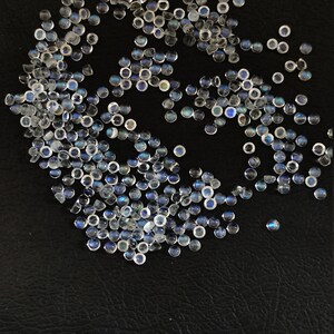 May include: A large group of iridescent white moonstone cabochons with a blue sheen, scattered on a black surface.