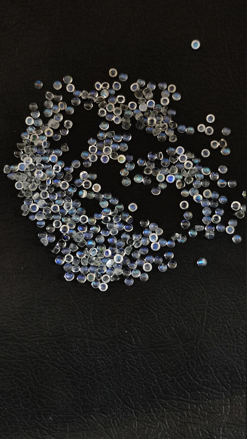 May include: A large group of iridescent white moonstone cabochons with a blue sheen, scattered on a black surface.
