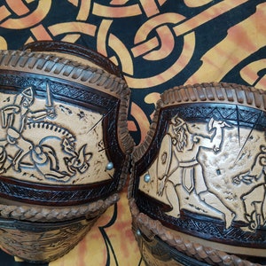 May include: Two leather arm guards with intricate carvings of Viking warriors. The guards are brown and tan with a braided leather trim.