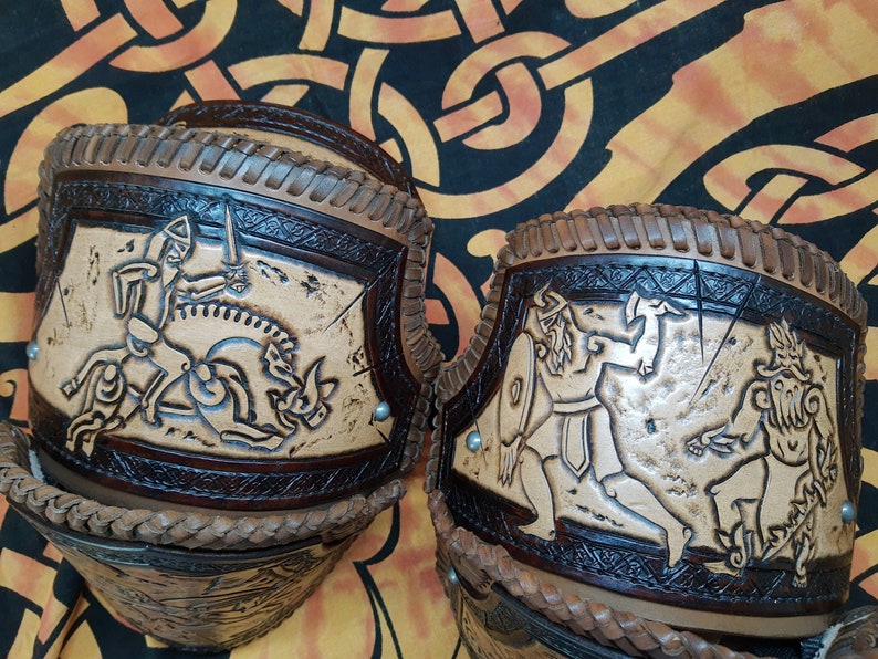 May include: Two leather arm guards with intricate carvings of Viking warriors. The guards are brown and tan with a braided leather trim.