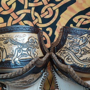 May include: Two leather leg guards with intricate carvings. One guard features a chariot scene with a horse and rider, while the other depicts a dragon. Both guards have a brown leather trim and are decorated with small metal studs.