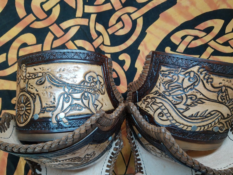 May include: Two leather leg guards with intricate carvings. One guard features a chariot scene with a horse and rider, while the other depicts a dragon. Both guards have a brown leather trim and are decorated with small metal studs.