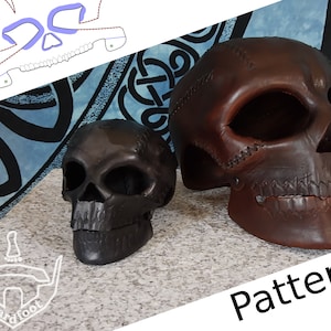 May include: Two leather skull sculptures, one brown and one black, with stitched details. The larger brown skull is on a white surface with the word "Pattern" in white text. The smaller black skull is on the left side of the image.