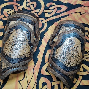 May include: A pair of brown leather arm guards with intricate carvings. One guard features a Viking ship with a dragon head, while the other depicts a mythical creature with wings.