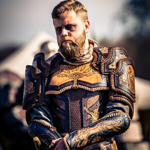May include: A man wearing a leather armor with intricate carvings and designs. The armor is brown and black with gold accents. The man has a beard and is looking directly at the camera.