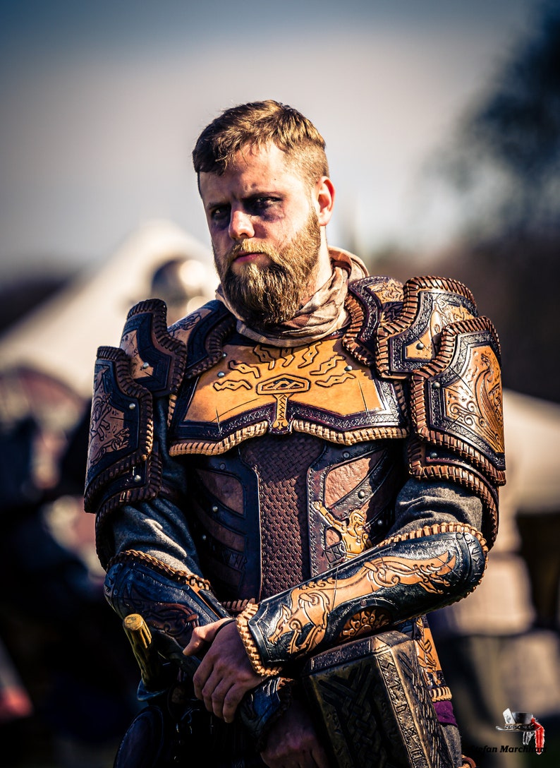 May include: A man wearing a leather armor with intricate carvings and designs. The armor is brown and black with gold accents. The man has a beard and is looking directly at the camera.