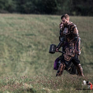 May include: A man in a leather armor suit with a large hammer in his hand walks through a grassy field. He is wearing a dark purple and black tunic and has a beard.