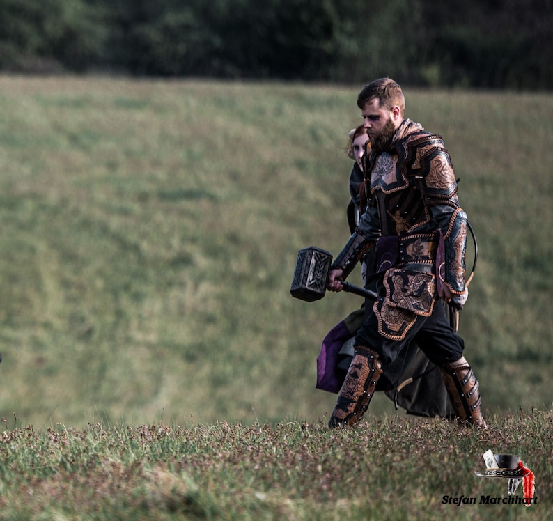 May include: A man in a leather armor suit with a large hammer in his hand walks through a grassy field. He is wearing a dark purple and black tunic and has a beard.