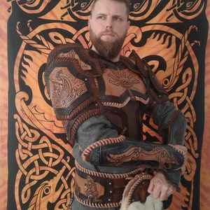 May include: A person wearing a brown leather armor with intricate carvings and a grey shirt. The armor has a belt with multiple straps and a pouch with white feathers hanging from it. The person is standing in front of a tapestry with a Celtic knot design.