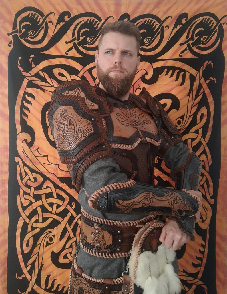 May include: A person wearing a brown leather armor with intricate carvings and a grey shirt. The armor has a belt with multiple straps and a pouch with white feathers hanging from it. The person is standing in front of a tapestry with a Celtic knot design.