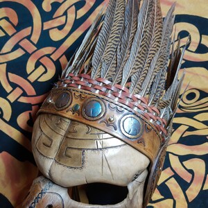 Pode incluir: Um capacete de couro com uma penacho de penas. O capacete &eacute; decorado com entalhes intrincados e detalhes met&aacute;licos. O penacho &eacute; feito de penas marrons e tem uma faixa de couro com detalhes met&aacute;licos.