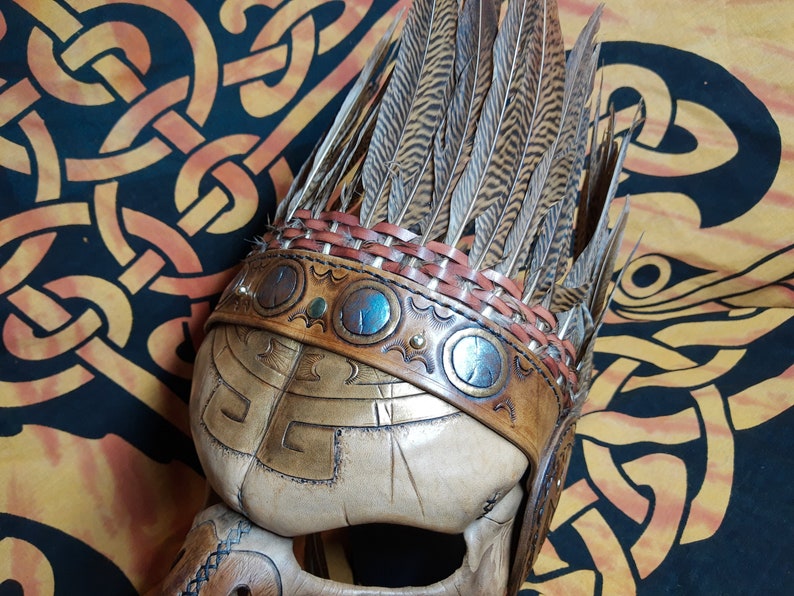 Pode incluir: Um capacete de couro com uma penacho de penas. O capacete &eacute; decorado com entalhes intrincados e detalhes met&aacute;licos. O penacho &eacute; feito de penas marrons e tem uma faixa de couro com detalhes met&aacute;licos.