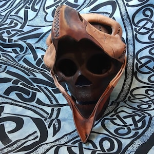 Pode incluir: Uma m&aacute;scara de caveira de couro marrom com uma capucha pontiaguda. A m&aacute;scara &eacute; feita de peda&ccedil;os de couro costurados e tem um design de caveira detalhado.