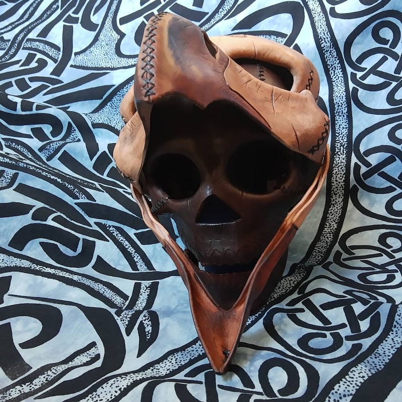 Pode incluir: Uma m&aacute;scara de caveira de couro marrom com uma capucha pontiaguda. A m&aacute;scara &eacute; feita de peda&ccedil;os de couro costurados e tem um design de caveira detalhado.