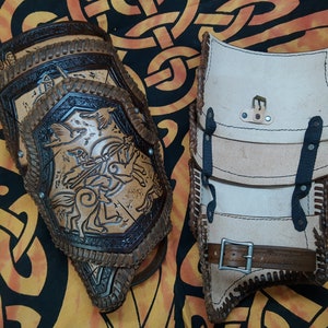 May include: Two leather arm guards, one brown with a detailed design and the other white with black straps. Both have decorative stitching.