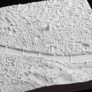 May include: A detailed, white 3D relief map of a city, showcasing intricate building and street layouts. The map includes a river and bridges, with a textured surface. The overall design is a monochromatic, architectural representation.