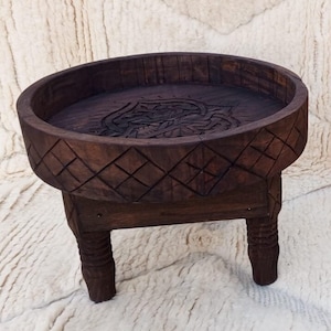 May include: A dark brown, hand-carved wooden table with a round top and three legs. The top features a carved floral design and is surrounded by a diamond pattern. The legs are carved with a ribbed design. The table is a decorative accent piece.