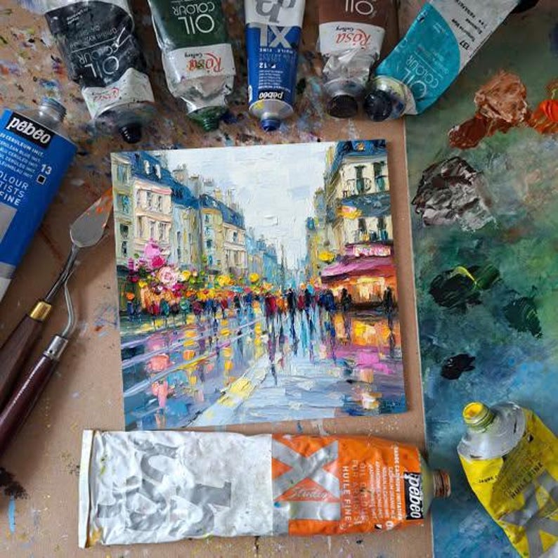 May include: An oil painting of a European city street scene with figures, buildings, and reflections. The painting is surrounded by tubes of oil paint and painting tools. The color palette includes blues, yellows, and oranges.