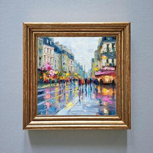 May include: Oil painting of a city street scene in a gold frame. The artwork depicts a wet street reflecting the lights of buildings and pedestrians. Buildings are painted in shades of blue, yellow, and pink.