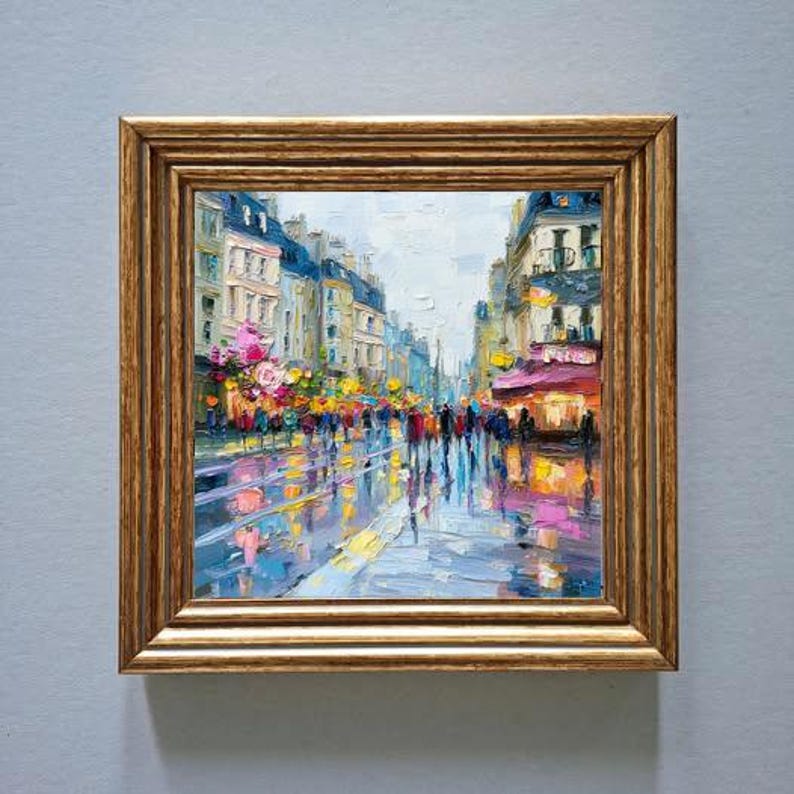 May include: Oil painting of a city street scene in a gold frame. The artwork depicts a wet street reflecting the lights of buildings and pedestrians. Buildings are painted in shades of blue, yellow, and pink.